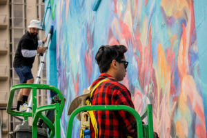 Aidan Kenny and Chris Perez on a ladder and lift to paint a large, colourful mural in Burlington