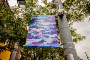 Close-up photograph of Hyedie Hashimoto's street light banner. It is painted in water colour in shades of purple and blue with dragonflies.