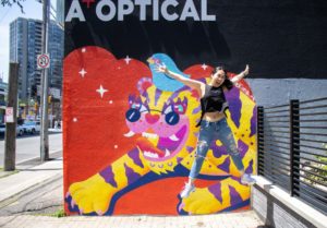 Jieun June Kim (artist) jumping in the air and smiling, in front of her new mural of a tiger and a bird