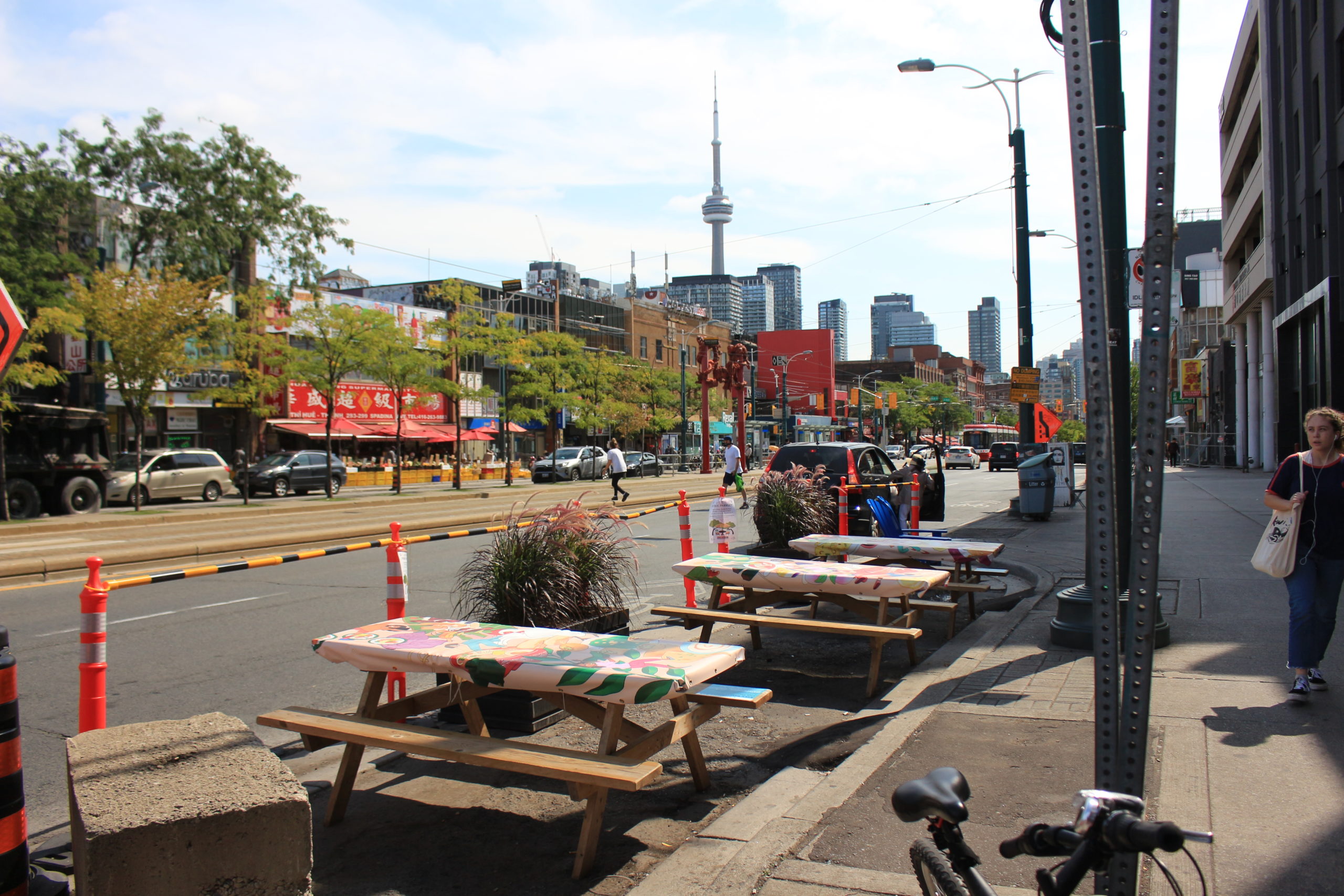 Photo of picnic tables with printed artwork on the sidewalk of Spadina Ave, storefronts and the CN tower in the distance