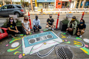 Six people wearing face masks and kneeling behind a ground mural by Romana Kassam of a cassette tape with the words HOW IT SHOULD BE
