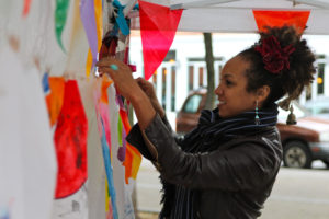Anna Jane McIntyre hanging colourful flags on a wall. She is Trinidadian and British and is wearing a leather jacket, jewelery accessories, with her hair tied up.