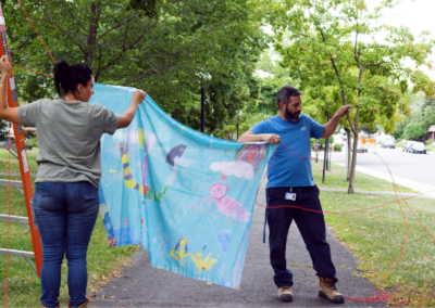 Photo of two people putting up Anna Jane McIntyre's banner that is sky blue with drawings. It is in a park with a path and green trees.