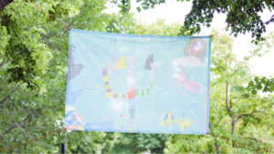 Photo of Anna Jane McIntyre's Banner installation, it is blue with colour children's drawings, the background is filled with green trees and an overcast sky.