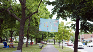Photo of Anna Jane McIntyre's Banner installation, it is blue with colour children's drawings, the background is filled with green trees and an overcast sky, and grey path.