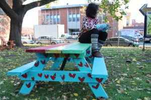A picnic table painted in blue, red, yellow and green with red hearts. Artist Anna Jane McIntyre is sitting on the bench and looking away