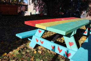 Close-up of a painted picnic table on a sunny day. The tabletop is painted red, yellow and green, and the rest of the furniture is blue with red hearts