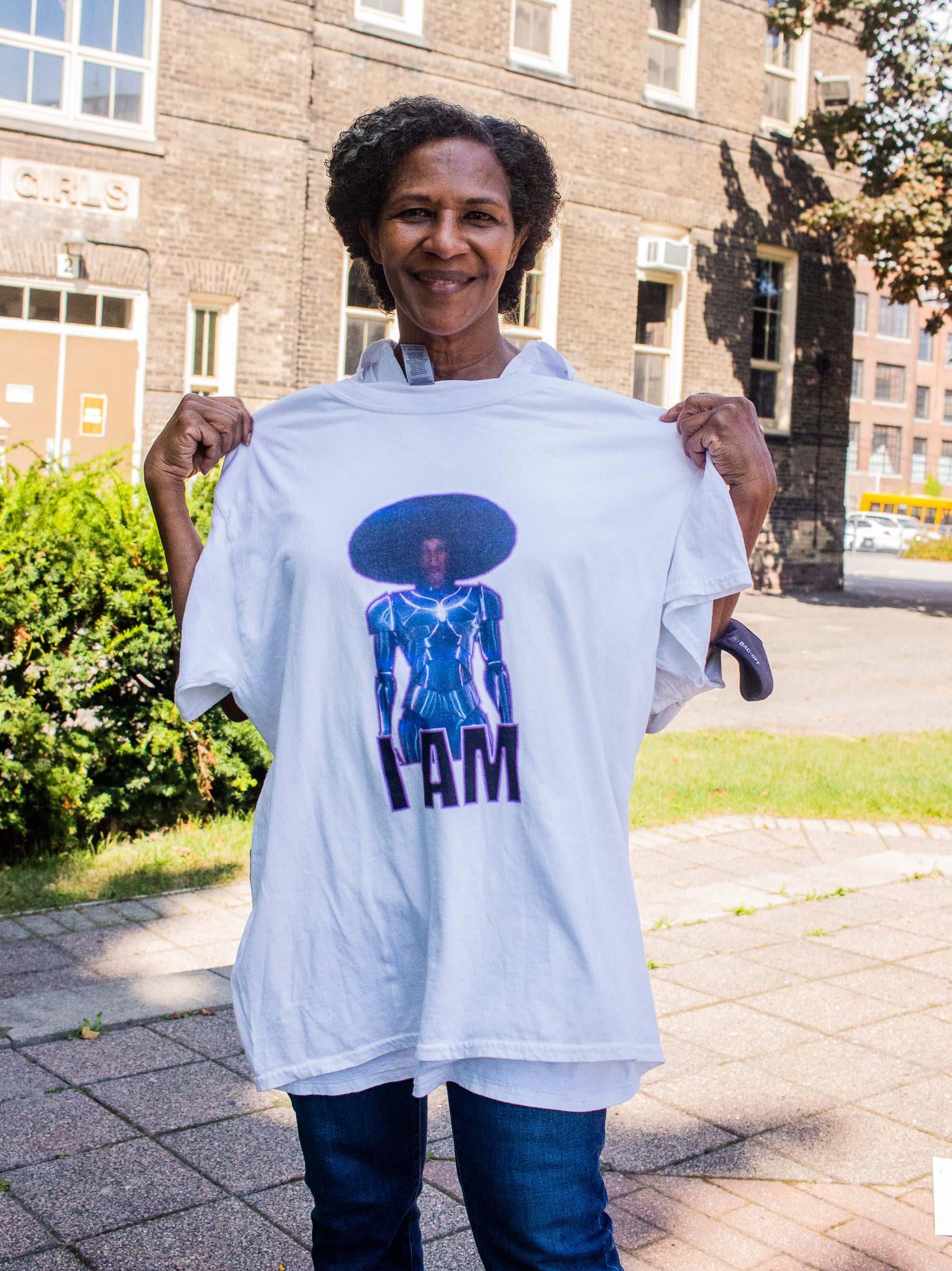 photo of Charmaine Lurch holding up a white tee with a graphic design of a woman. She is smiling and standing in front of a building and trees