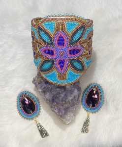 Photo of beaded floral cuff and earrings by artist Lindsey Lickers, its displayed on a rock and has colours of teal, pink, purple and orange.
