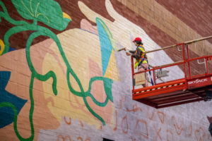 Artist Wenting Li on a scissor lift and painting a mural on a large wall in Toronto's Chinatown