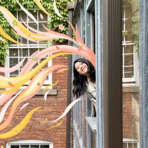Artist Meegan Lim poking her head out of a building window and smiling. There are drawn graphics on the photo of peach and orange waves coming out of the building.