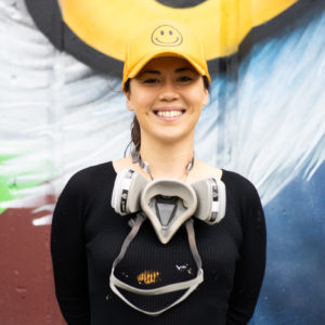 Photo of mural assistant Kseniya Tsoy, she is smiling at the camera, wearing a yellow hat, black shirt, and a grey painting mask. She is in front of a mural that has an owl eye with colours of blue, yellow, purple, green and burgundy.