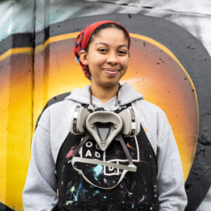 Photo of mural assistant Edan Maxam, smiling at the camera, wearing a grey hoodie and black apron with OCAD U logo on it. There is a spray paint mask around her neck, with paint splatters on her clothes. Edan is standing in front of a mural with colours of black, yellow and grey.