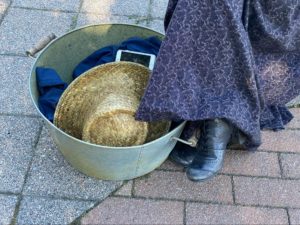 Photo of a metal bin with a hat, blue cloth and phone. Charmaine Lurch's leg is cropped showing one of her boots and skirt - standing on paving stones.