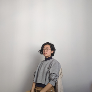 Florence Yee standing against a white wall. They are of East Asian descent with short dark hair, glasses, and wearing a grey sweater.
