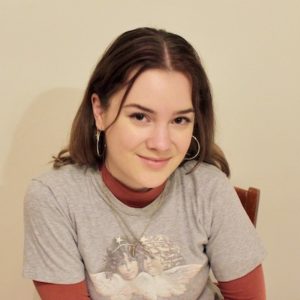 Eva Morrison smiling at the camera. Eva is sitting on a chair and has long brown hair. She is wearing a red long-sleeve underneath a grey t-shirt.