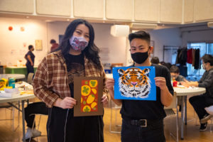 Artists Meegan Lim (left) and Paddy Leung (right) holding up examples of their artwork at a workshop at Cecil Community Centre, there are tables with people at them in the background.
