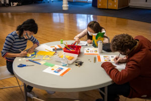 Photo of a family creating artwork around a round grey table in Cecil community centre. There are art supplies, instructions sheets and scissors on the table.
