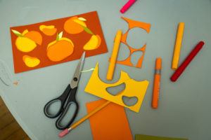 Photo of paper cut art with markers, yellow and orange cardstock, scissors on a grey table.