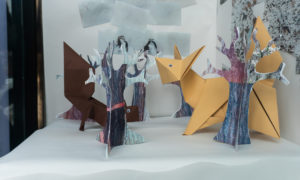 Close up photo of an paper art installation in a window display, it has paper trees, snow and animals.