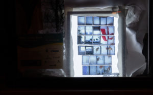 Photo of an art installation with a lightbox showing photos of someones home, there are paper cutouts surrounding it.