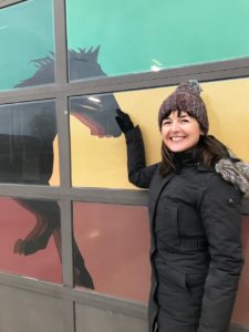 Vertical photo of artist Pam Lostracco in front of her artwork on the Brampton Fire Station. It features colours of blue, green, yellow and red with a silhouette of a horse.