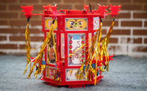 Photo of red Yue Moon lantern with artwork framed and golden tinsel hanging from the corners. It is sitting on the ground, next to a brick wall.