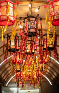 Vertical photo of 20 palace lanterns in red and black acrylic holding community artwork for Yue Moon. The lanterns have red and gold tinsel hanging from the corners. They are hanging in an arched pathway at night.