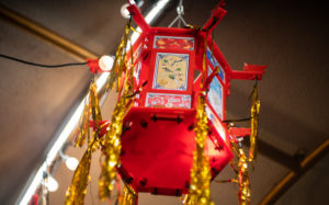 ID2: Horizontal photo of a red palace lantern that holds artwork featuring images of food and Lunar New Year with gold tinsel hanging from the corners. The lantern is hanging from an archway at night with string lights.