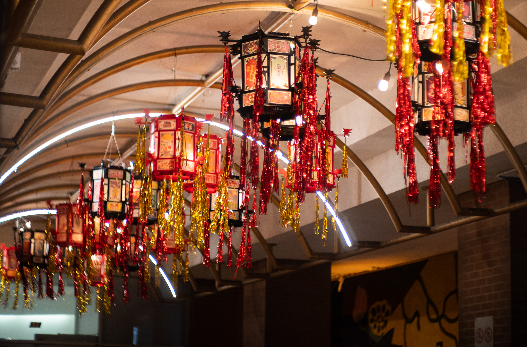 ID3: Horizontal photo of 20 palace lanterns in red and black acrylic holding community artwork for Yue Moon. The lanterns have red and gold tinsel hanging from the corners. They are hanging in an arched pathway at night.