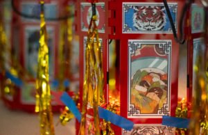 Photo of a Chinese palace lantern with red borders and artwork on each panel. There is gold tinsel and illumination within each lantern.