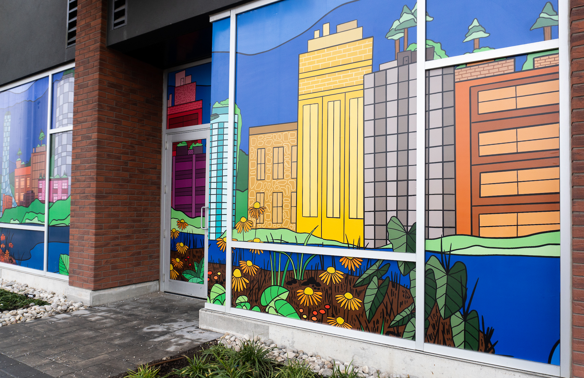 A storefront window with large murals covering the windows. It has colourful and bold imagery of city buildings, nature, grassy hills, and blue skies representing community, growth and convergence