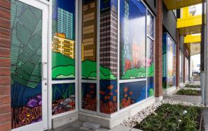 A side angle view of a series of storefront window murals. The windows are covered in colourful illustrations with bold outlines of a city with nature, grassy hills, water and blue skies