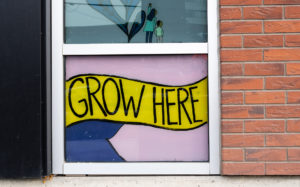 A close-up of a window on the side of a building that is painted with a pink and navy background with the words GROW HERE in a yellow ribbon