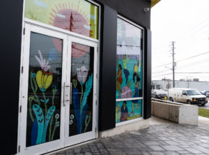 A storefront with the windows and glass doors painted with a colourful mural. Imagery showcases nature, flowers, and community to represent growth and community.