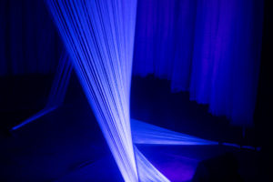 A close-up of a multimedia light installation in a window storefront at night. It is glowing blue purple light with strands of white thread creating illusions from ceiling to ground.