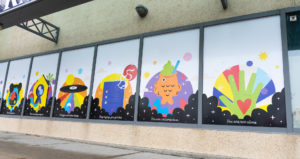 A building with large window panels that are filled with large scale mural prints with motivational quotes and colourful graphics of shapes, rainbows, and different fun icons representing happiness and community