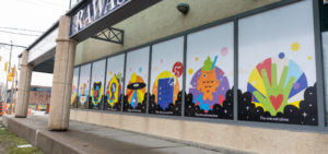 A building with large window panels that are filled with large scale mural prints with motivational quotes and colourful graphics of shapes, rainbows, and different fun icons representing happiness and community