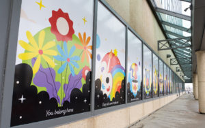 Close-up of a building with lots of panels along the side with large mural prints. The prints have motivational quotes and colourful graphics of flowers, rainbows, and other symbols of community and family