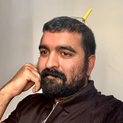 Headshot of artist Arjun Lal, a man of East Asian descent with dark short hair, moustache and beard. His head is resting on his hand and gazing off camera with a head massager on the back of his head