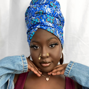 Headshot of artist Laura O. Okos-Iboje posing in front of a white background. The artist is posing with her hands under her chin and is wearing a bright blue printed headdress, large gold hoops in her ears and gold nose rings and a necklace.