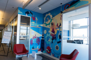 Photograph of an interior mural installed in an office space, with chairs in front of it and glass doors around it. The mural features imagery of buildings, gears, silhouetted figures and other details set against a bright blue background.