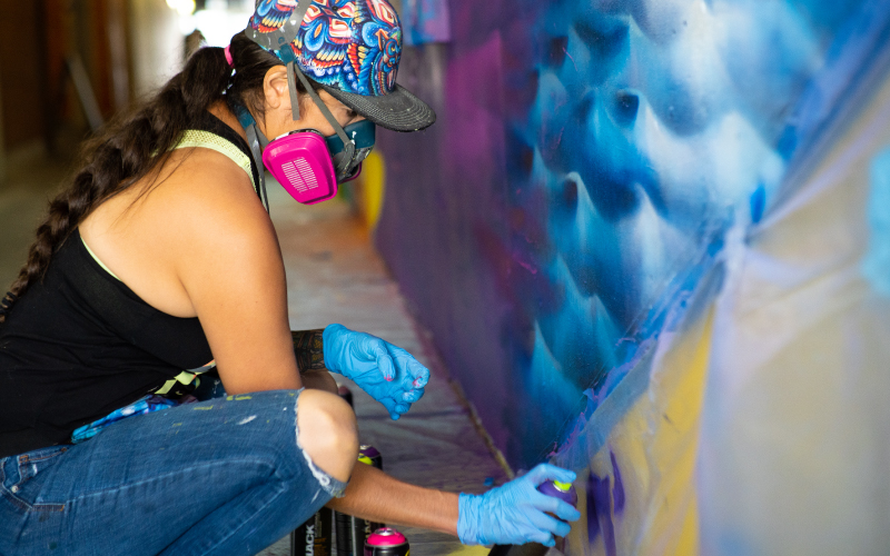 Artist Shalak Attack in PPE and spray painting a mural as part of the Daily Migration Mural project with STEPS Public Art