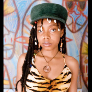 Headshot of artist Ehiko Odeh who has dark skin and long braids. She is wearing a tiger print tank top and a dark green hat in front of a colourful backdrop
