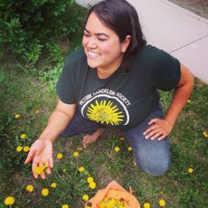 A candid of Latifa Pelletier-Ahmed who is kneeling on grass with dandelions growing. SHe has dark hair and wearing a t-shirt with a flower on it
