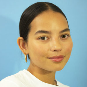 Headshot of artist Tayler Buss. Tayler is wearing gold hoop earrings with hair slicked back and standing against a light blue backdrop.
