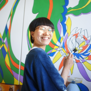 Headshot of artist Elaine Victoria Wang posing in front of a large painting that they are painting. She is turned towards the camera and is smiling while wearing a blue top.