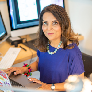 Headshot of artist Farida Zaman sitting at a desk and surrounded by monitors. She is wearing a blue top and wearing a colourful necklace and bracelet as she looks up into the camera.