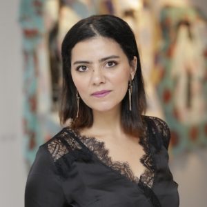 Headshot of Ichraq Bouzidi (she/her) who has a tanned complexion and straight medium length hair. She is wearing a black blouse and smiling at the camera
