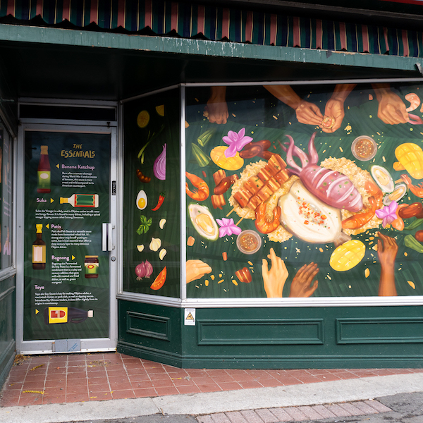 A photograph of a storefront window and door covered with illustrations of food. It features imagery of hands reaching out to partake in the wide variety of food drawn in the centre, set against a dark green background. 